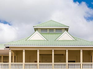Standing-Seam-Metal-Roof-Zanesville-OH-Ohio-2 | Cosam Contracting Beige building with a green metal roof, triangular dormers, and a covered wraparound porch with columns.